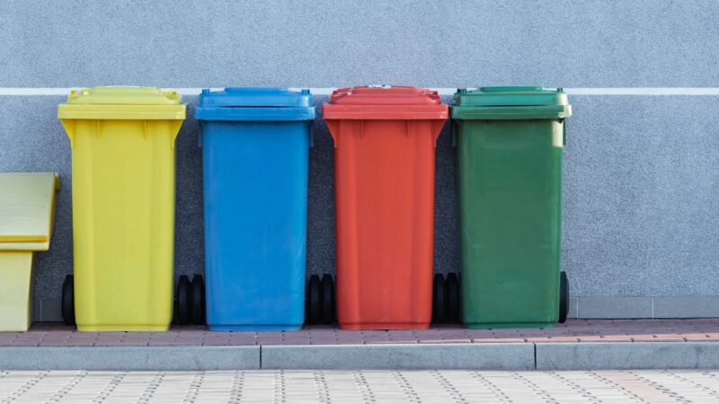 A row of multicolored recycling bins against a gray wall.