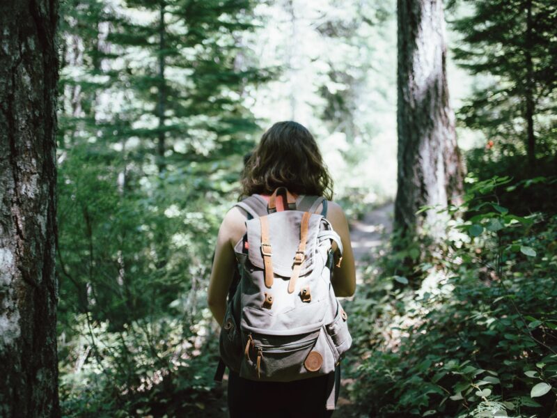 A female hiker with dark hair stands amidst out-of-focus trees, looking away from the camera and wearing a backpack.